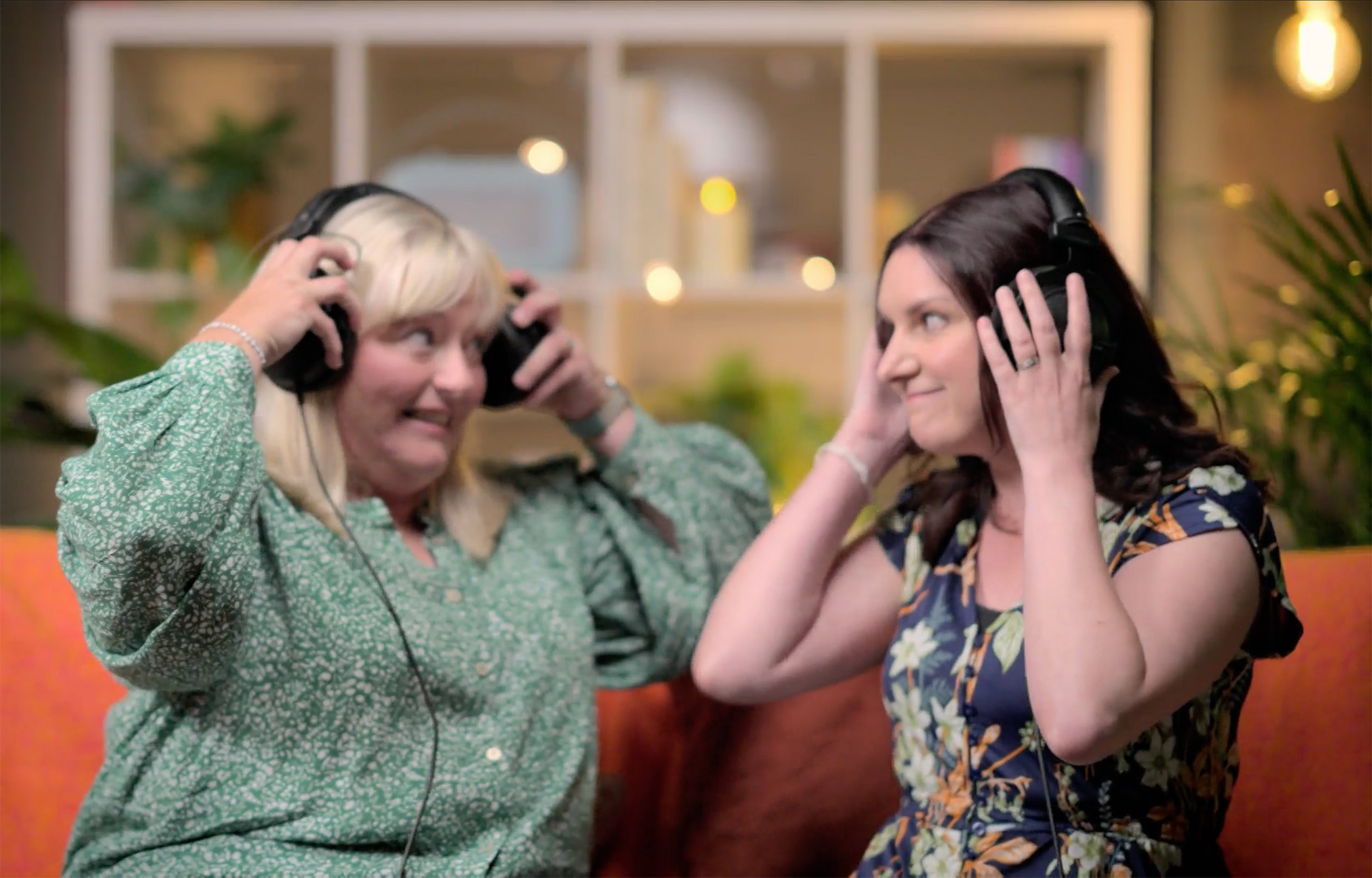 Two women sitting on a sofa putting on headphones and smiling